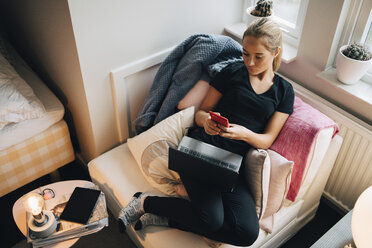 Blick von oben auf ein Teenager-Mädchen, das ein Smartphone benutzt, während es sich mit seinem Laptop auf dem Sitz zu Hause zurücklehnt - MASF00940