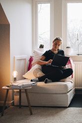 Teenager-Mädchen in voller Länge mit Laptop auf dem Sitz gegen das Fenster zu Hause liegend - MASF00943