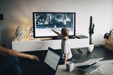 Kniender Junge in voller Länge beim Berühren eines Smart-TVs im heimischen Wohnzimmer - MASF00945