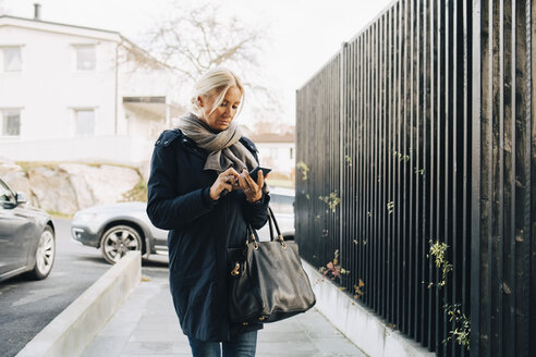 Ältere Frau, die ihr Smartphone benutzt, während sie auf dem Fußweg am Zaun spazieren geht - MASF00953