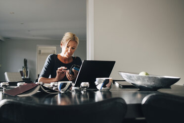 Reife blonde Frau mit Smartphone und Laptop auf dem Esstisch zu Hause - MASF00961