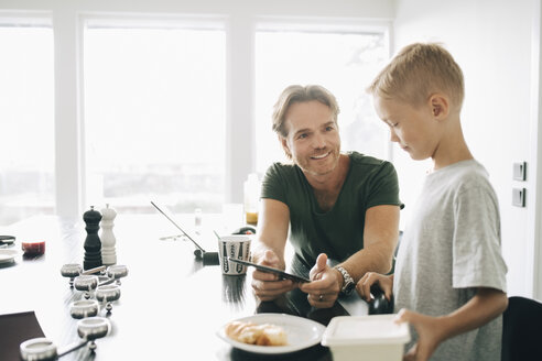 Ein lächelnder Mann zeigt einem Jungen am Esstisch vor dem Fenster ein digitales Tablet - MASF00965