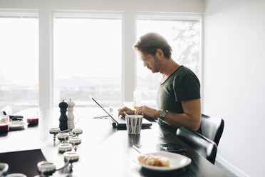 Side view of mature man sitting at dining table using laptop against window - MASF00967