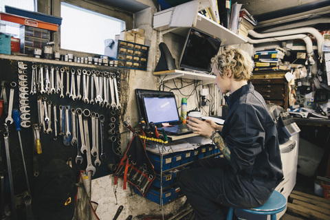 Seitenansicht einer Mechanikerin, die einen Einwegbecher hält, während sie einen Laptop in einer Autowerkstatt benutzt, lizenzfreies Stockfoto