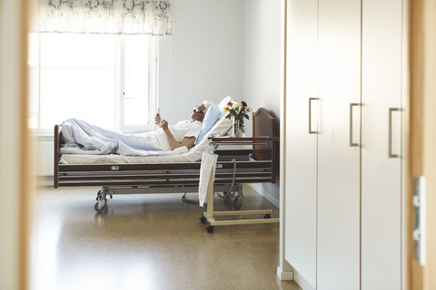 Seitenansicht eines älteren Mannes, der ein Smartphone benutzt, während er auf dem Bett in einer Krankenstation liegt, lizenzfreies Stockfoto