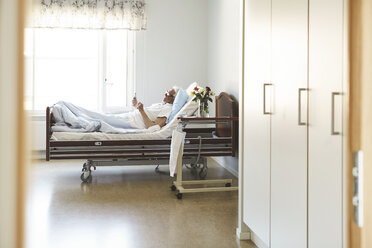 Seitenansicht eines älteren Mannes, der ein Smartphone benutzt, während er auf dem Bett in einer Krankenstation liegt - MASF01007
