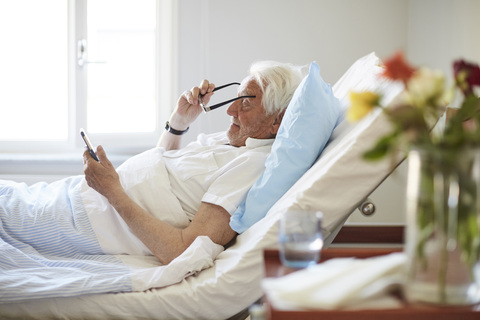 Seitenansicht eines älteren Mannes, der eine Brille trägt, während er ein Smartphone in einer Krankenstation benutzt, lizenzfreies Stockfoto