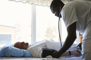 Seitenansicht eines Krankenpflegers, der den Blutdruck eines älteren Mannes auf einem Krankenhausbett vor einem Fenster überprüft - MASF01013