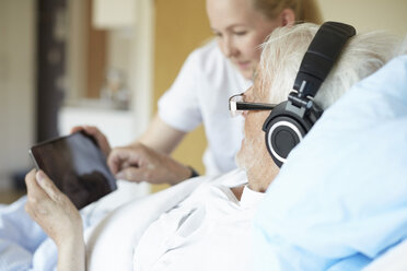 Senior man wearing headphones while using digital tablet with female nurse on hospital bed - MASF01019