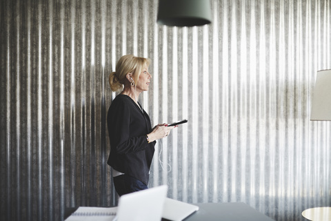 Seitenansicht einer Geschäftsfrau, die mit einem Mobiltelefon an einer Wellblechwand in einem Kreativbüro spricht, lizenzfreies Stockfoto