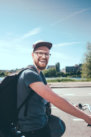 Seitenansicht Porträt eines glücklichen Mannes, der einen Rucksack trägt, während er mit dem Fahrrad auf der Straße gegen den Himmel fährt, lizenzfreies Stockfoto