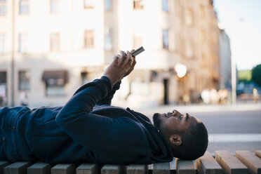 Seitenansicht eines Mannes, der auf einer Bank liegt und sein Smartphone in der Stadt benutzt - MASF01041