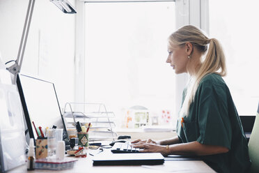 Seitenansicht einer Krankenschwester, die an einem Computertisch vor einem Fenster im Büro arbeitet - MASF01046