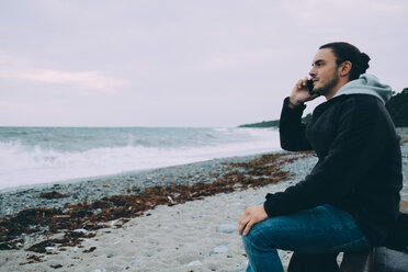 Seitenansicht eines jungen Mannes, der am Strand sitzend ein Mobiltelefon benutzt - MASF01049