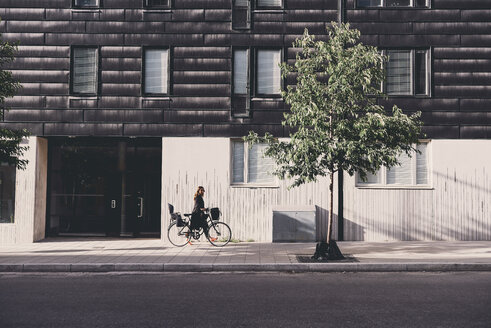 Seitenansicht der reifen Frau zu Fuß mit Fahrrad auf dem Bürgersteig gegen Gebäude in der Stadt - MASF01064
