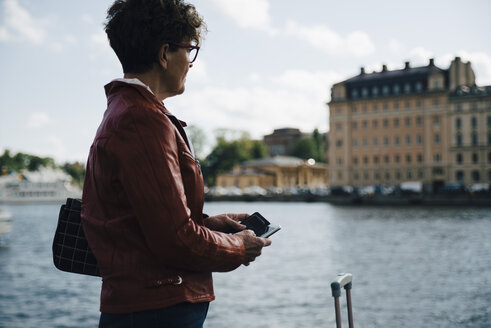 Seitenansicht einer älteren Frau, die ihr Handy in der Hand hält, während sie in der Stadt am Meer steht - MASF01087