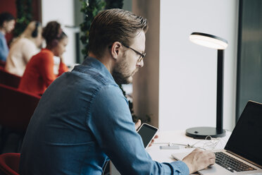 Seitenansicht eines Geschäftsmannes, der einen Laptop benutzt, während er mit seinen Kollegen im Büro am Schreibtisch sitzt - MASF01093
