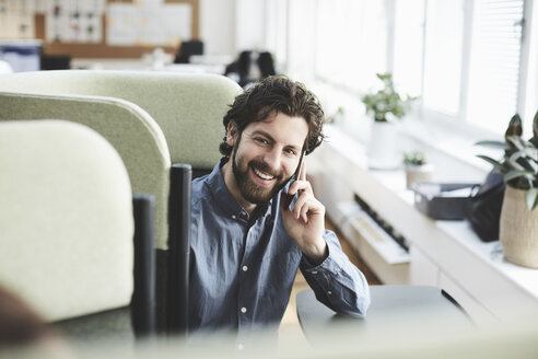 Lächelnde männliche Fachkraft, die über ihr Mobiltelefon spricht, während sie auf einem Stuhl im Kreativbüro sitzt - MASF01128