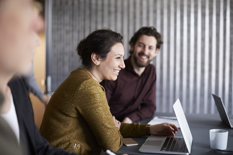 Lächelndes, kreatives Geschäftsteam sitzt mit Laptop am Konferenztisch im Sitzungssaal, lizenzfreies Stockfoto