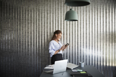 Lächelnde reife Geschäftsfrau, die ein Mobiltelefon benutzt, während sie an einer Wellblechwand im Kreativbüro steht - MASF01140