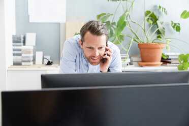 Smiling businessman talking on mobile phone while using computer at creative office - MASF01151