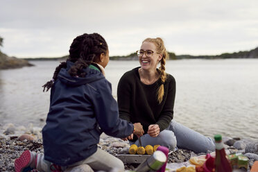 Lächelnde Frau und Tochter kochen Mais auf dem Grill am Strand bei Sonnenuntergang - MASF01159
