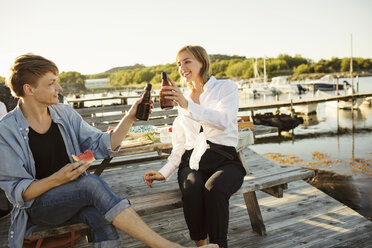 Smiling male and female friends toasting beer bottles at harbor - MASF01166