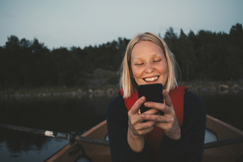 Lächelnde Frau, die ein Mobiltelefon benutzt, während sie in einem Boot auf einem See sitzt, lizenzfreies Stockfoto