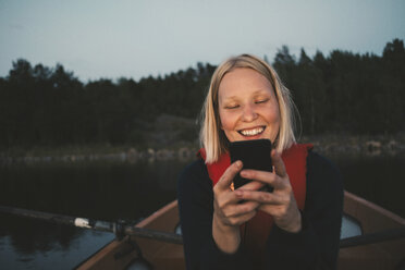 Lächelnde Frau, die ein Mobiltelefon benutzt, während sie in einem Boot auf einem See sitzt - MASF01179