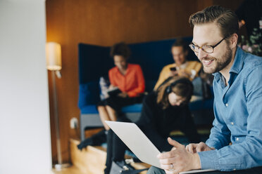 Smiling mid adult businessman using laptop while sitting with colleagues at office in cafeteria - MASF01201