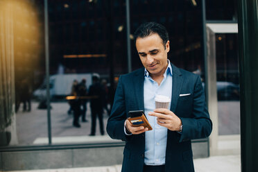 Lächelnder Geschäftsmann, der sein Smartphone benutzt und einen Einweg-Kaffeebecher gegen ein Gebäude in der Stadt hält - MASF01223