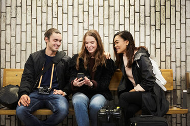 Smiling young friends looking at smart phone while sitting against wall on subway station platform - MASF01231
