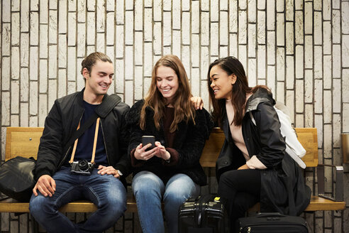 Lächelnde junge Freunde schauen auf ihr Smartphone, während sie auf dem Bahnsteig einer U-Bahn-Station an der Wand sitzen - MASF01231