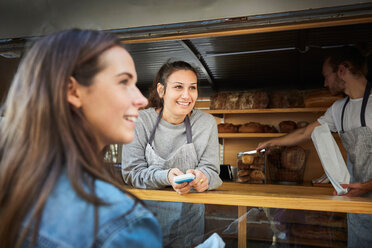 Lächelnde Verkäuferin, die die Zahlung von einem Kunden annimmt, während ihr Kollege am Imbissstand Brot verpackt - MASF01250