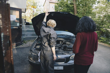 Rückansicht eines Kunden, der bei einer Mechanikerin vor einer Autowerkstatt steht - MASF01274