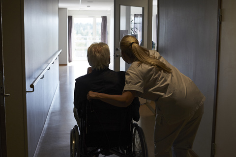 Rückansicht einer Krankenschwester mit einem älteren Mann im Rollstuhl auf dem Krankenhausflur, lizenzfreies Stockfoto