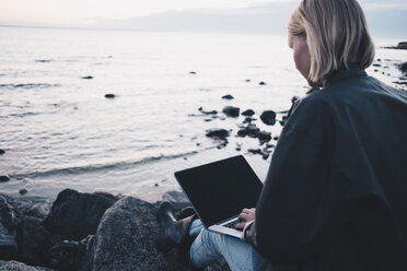 Rückansicht einer Frau, die einen Laptop benutzt, während sie auf einem Felsen am Seeufer sitzt - MASF01308