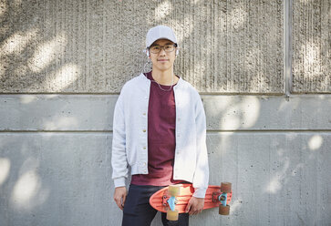 Porträt eines jungen Mannes mit Skateboard, der an einer Wand auf dem Universitätsgelände steht - MASF01316