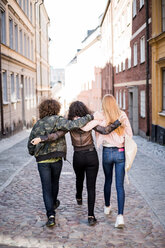 Rear view of friends walking with arms around on cobbled street in city - MASF01319
