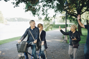 Senior couple waving friends riding tandem bike in park - MASF01332