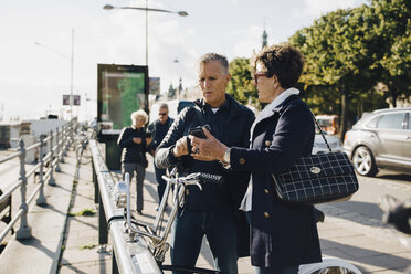 Senior couple using mobile phone while standing on sidewalk in city - MASF01333