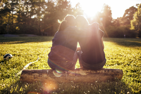 Rückansicht von zärtlichen Schwestern, die an einem sonnigen Tag im Park auf einem Baumstamm sitzen, lizenzfreies Stockfoto