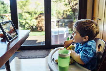 Boy looking at digital tablet and eating food at home - CAVF35174