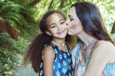 Mother kissing daughter on cheeks against trees - CAVF35214