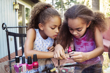 Mädchen trägt Nagellack auf die Fingernägel ihrer Schwester auf, während sie an einem Tisch im Hof sitzt - CAVF35234