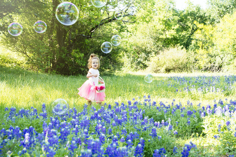 Lächelndes Mädchen, das sich Seifenblasen ansieht, während es neben blühenden Blumen auf einem Feld steht, lizenzfreies Stockfoto
