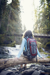 Young woman sitting on tree log in stream - CAVF35344