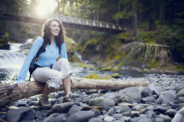 Young woman sitting on tree log in stream - CAVF35345