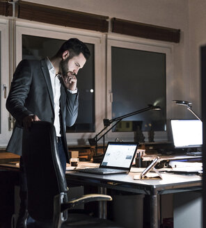 Geschäftsmann steht nachts im Büro und schaut auf einen Laptop - UUF13201