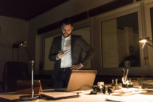 Geschäftsmann steht nachts im Büro und benutzt sein Smartphone - UUF13204